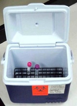 Biohazard-labeled cooler containing two test tubes with purple caps placed on a black grid, indicating transport of potentially hazardous biological samples